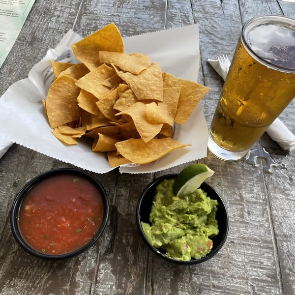 A basket of tortilla chips with salsa, guacamole, and a glass of beer on a wooden table.